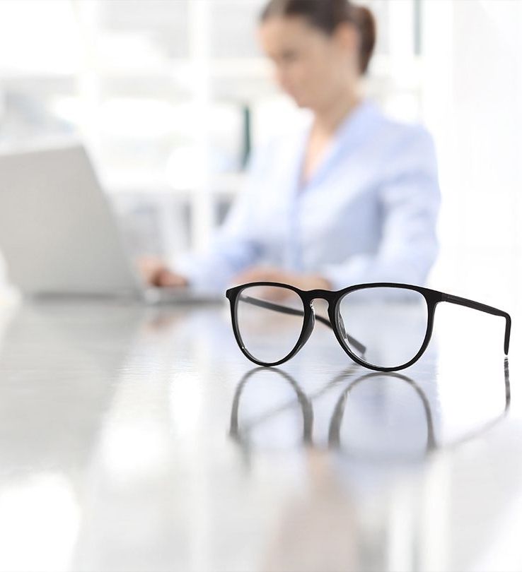 Eine Arbeitsplatzbrille im Vordergrund und im Hintergrund eine Person die am Laptop arbeitet